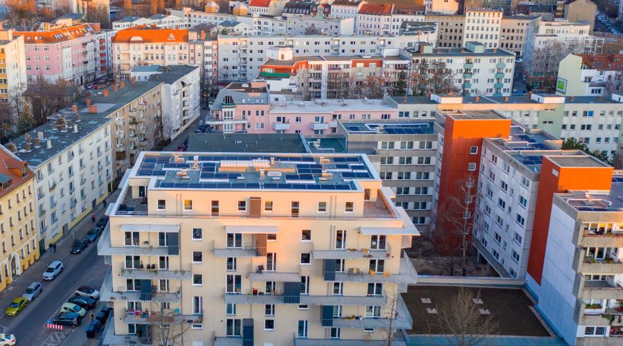 Luftaufnahme eines Stadtviertels mit modernen Wohngebäuden, einige mit Solarpaneelen auf den Dächern. Die Gebäude sind dicht bebaut, und die Sonne taucht die Szene in ein warmes Licht.