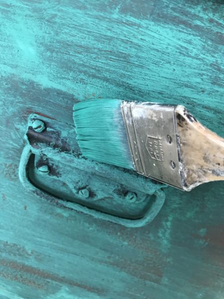 Nahaufnahme eines Pinsels mit türkisen Borsten, der Kreidefarbe auf eine strukturierte, türkise Oberfläche aufträgt. Ein alter, abgenutzter Metallgriff ist ebenfalls teilweise mit Farbe bedeckt, was den Shabby-Chic-Look unterstreicht.