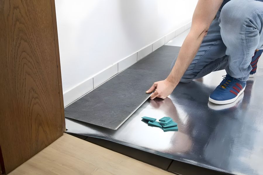 Eine Person in Jeans und blauen Sneakers verlegt dunkle Vinylbodenpaneele mit Klicksystem auf einer silbernen Trittschalldämmung. An der Wand sind weiße Fliesensockelleisten zu sehen. Im Vordergrund liegen grüne Abstandskeile zur Fixierung des Dehnungsabs