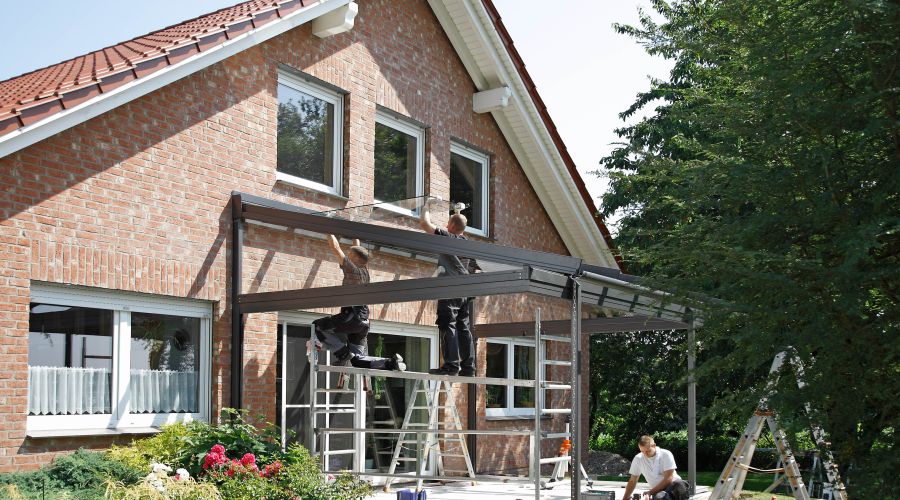 Handwerker montieren ein Glas-Terrassendach an einem roten Backsteinhaus. Zwei Arbeiter stehen auf einem Gerüst und befestigen Glasplatten an der Metallkonstruktion, während ein weiterer Arbeiter am Boden arbeitet. Im Vordergrund blühen rote Rosen in eine