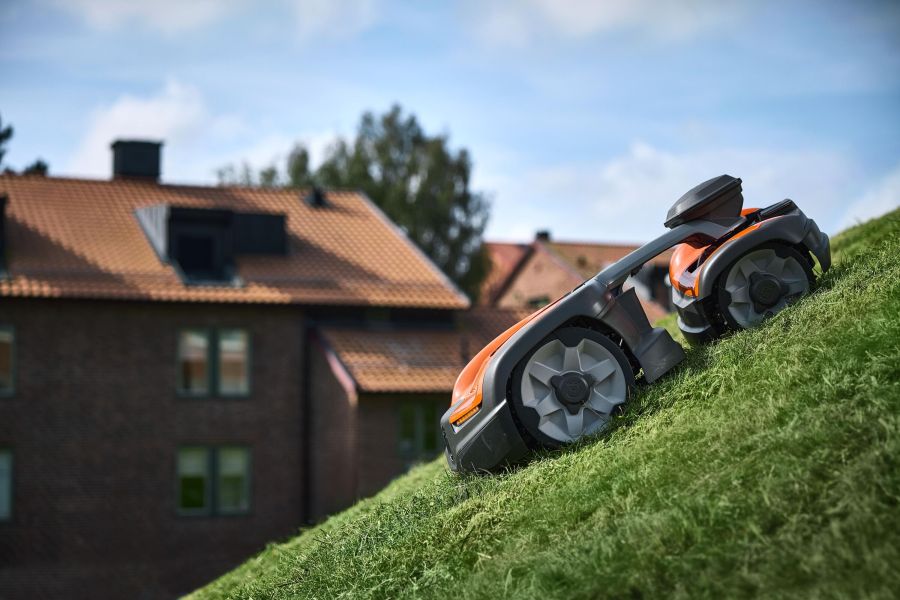 Auf dem Bild ist ein orangefarbener Rasenmähroboter auf einem grünen Hügel zu sehen. Im Hintergrund erheben sich Wohnhäuser mit roten Ziegeldächern unter einem hellblauen Himmel mit wenigen Wolken. Die Szene wirkt ruhig und modern, als würde die Gartenarb