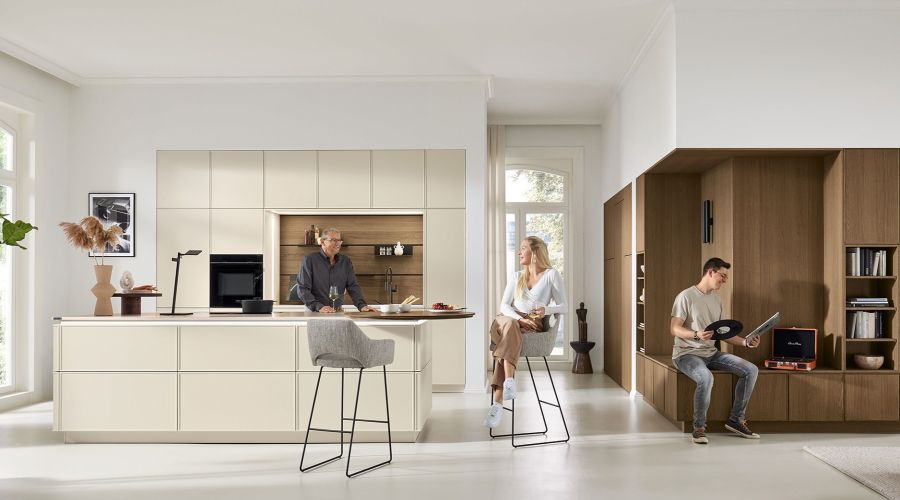Eine helle, moderne Küche mit einer großen Kochinsel in Beige und Holzoptik. Drei Personen beleben den Raum: Ein Mann steht an der Insel, eine Frau sitzt auf einem hohen Barhocker und ein junger Mann sitzt auf einer integrierten Holzbank.