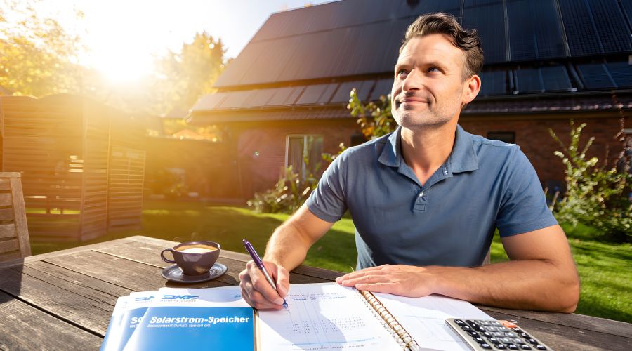 Ein Mann sitzt an einem sonnigen Gartentisch und macht sich mit Notizbuch und Taschenrechner an die Planung seiner Solaranlage. Im Hintergrund ist sein Haus mit Photovoltaik-Modulen auf dem Dach zu sehen.