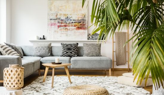 Ein helles, modernes Wohnzimmer mit einem großen grauen Ecksofa und verschiedenen schwarz-weiß gemusterten Kissen. Ein runder Jute-Pouf liegt auf einem hellen Teppich vor einem niedrigen Holztisch, während eine große Palme rechts den Raum mit Grün belebt.