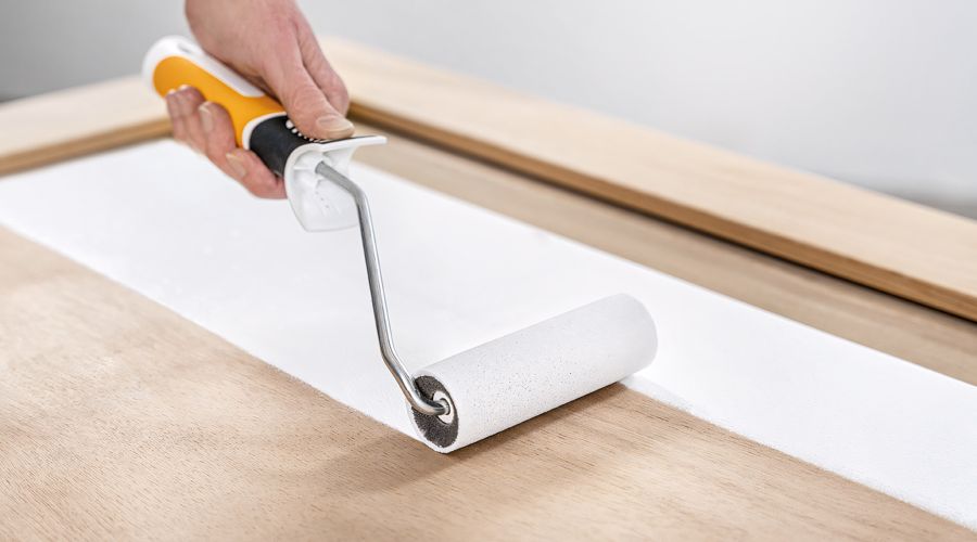 Eine Hand mit einem gelb-weißen Griff führt eine kleine Schaumstoff-Lackrolle über eine glatte, helle Holztür. Die Rolle hinterlässt eine saubere, deckende Bahn weißer Farbe auf dem unbehandelten Holz.