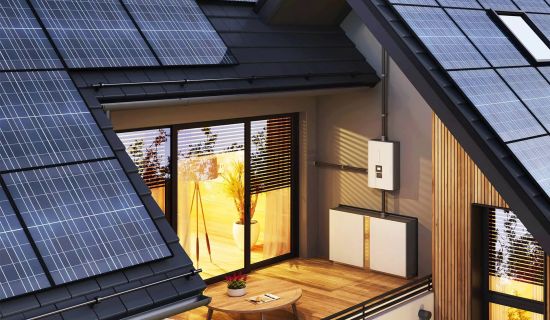 An der Außenwand eines modernen Hauses mit Solaranlage ist auf einem Holzbalkon ein weißer Batteriespeicher montiert.