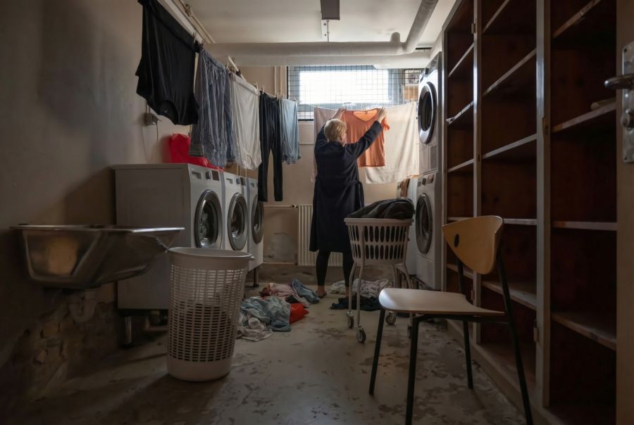 Ein funktionaler Kellerraum, der als Waschküche dient, mit mehreren weißen Waschmaschinen an der Wand. Eine Person steht mit dem Rücken zur Kamera und hängt ein orangefarbenes Kleidungsstück auf eine Wäscheleine, an der bereits andere Kleidung trocknet.