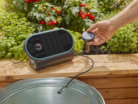 Ein solarbetriebenes Bewässerungsgerät ist am Rande eines Hochbeets platziert. Ein Ansaugschlauch führt von dem Gerät zu einer darunterstehenden Zinkwanne, während eine Hand einen Bodensensor in die Erde neben Tomaten- und Kräuterpflanzen steckt.