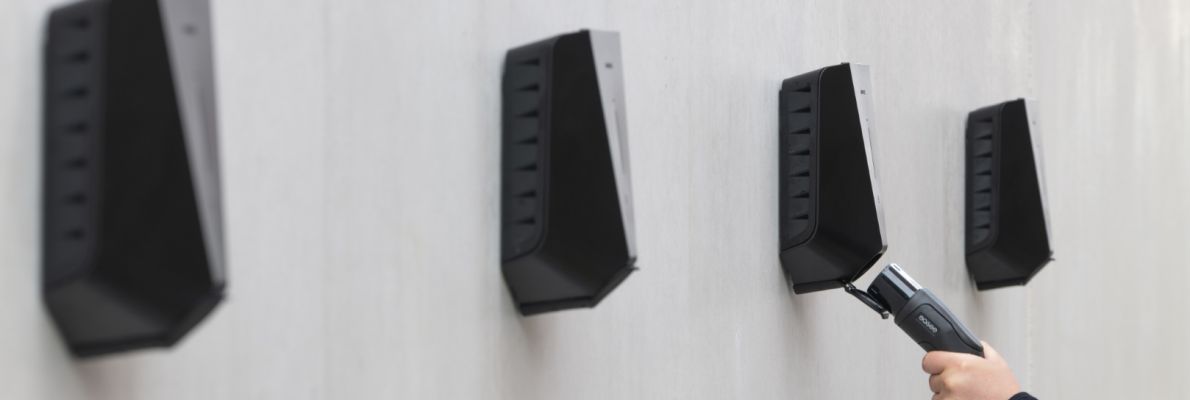 Vier moderne, schwarze Wallboxen sind in einer Reihe an einer schlichten Betonwand montiert. Die Hand einer Person führt von rechts ein Ladekabel in eine der Wallboxen ein, um den Ladevorgang zu starten.