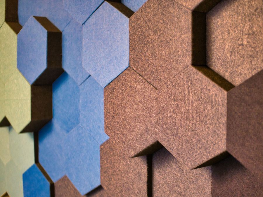 Dieses Bild zeigt eine Nahaufnahme einer Wandverkleidung aus dreidimensionalen, sechseckigen Akustikpaneelen in den Farben Blau, Grün und Braun. Die geometrische Struktur und die leicht raue Textur erzeugen eine moderne und abstrakte Atmosphäre.