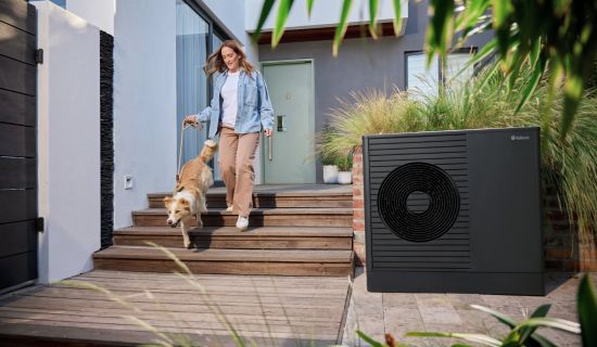 Eine Frau geht mit ihrem Hund eine flache Holztreppe vor einem modernen Einfamilienhaus hinunter. Rechts im Vordergrund steht eine kompakte, dunkelgraue Luft-Wasser-Wärmepumpe unauffällig im Außenbereich.