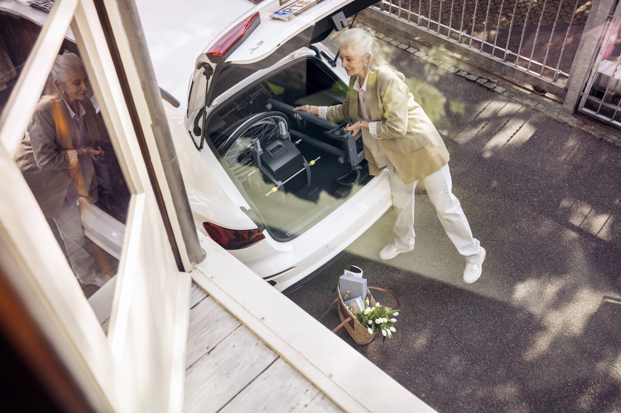 Ältere Frau verstaut einen zerlegten Rollstuhl und eine Treppensteighilfe im Kofferraum eines weißen Autos. Im Vordergrund steht eine Tasche mit Tulpen.