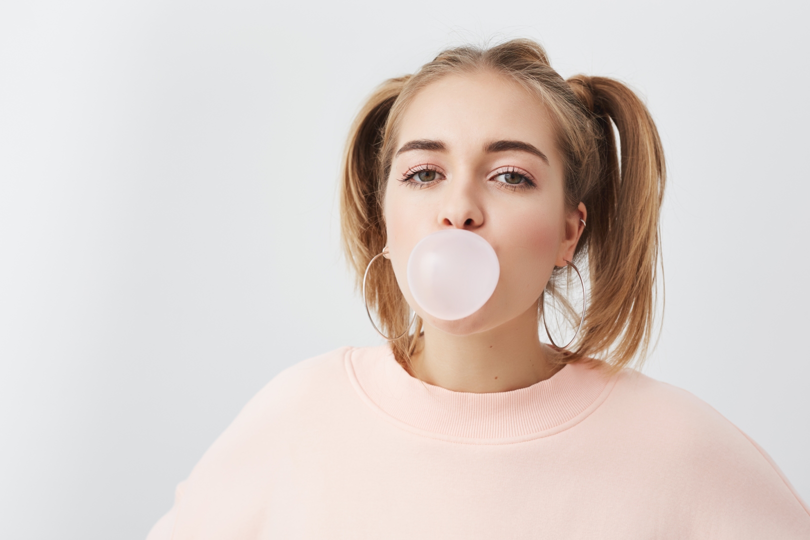 Ein junges, blondes Mädchen mit zwei Pferdeschwänzen bläst eine große rosa Blase aus Kaugummi. Sie trägt ein rosa Sweatshirt und große, runde Ohrringe. Das Bild steht symbolisch für knifflige Flecken wie Kaugummi.