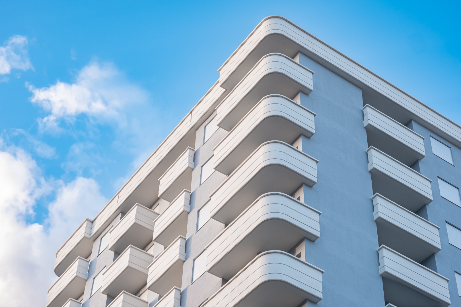 Die Fassade eines modernen Mehrfamilienhauses mit grauer Verkleidung und weißen, abgerundeten Balkonen, fotografiert von unten gegen einen blauen Himmel. Das Bild steht symbolisch für Mietobjekte, in denen die Trinkwasserverordnung eine wichtige Rolle spi