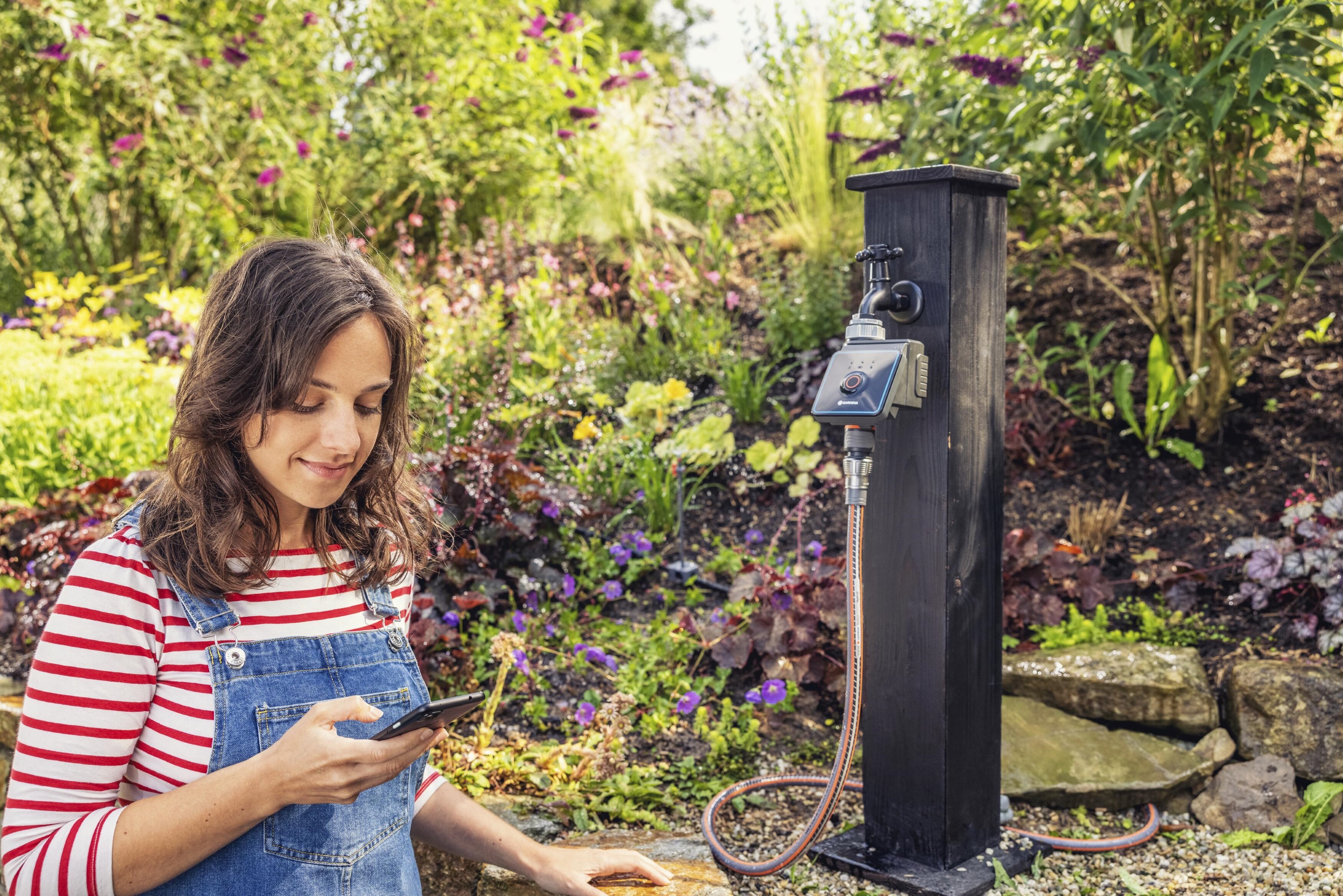 Eine Frau sitzt in einem bunten Garten und schaut auf ihr Smartphone. Daneben ist ein Wasseranschluss mit digitalem Bewässerungscomputer zu sehen.